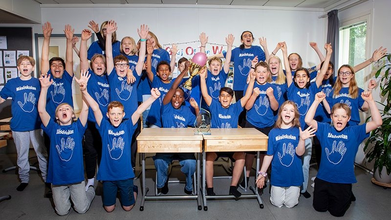 Klass med elever i blå t-shirts som sträcker upp händerna i luften i sitt klassrum